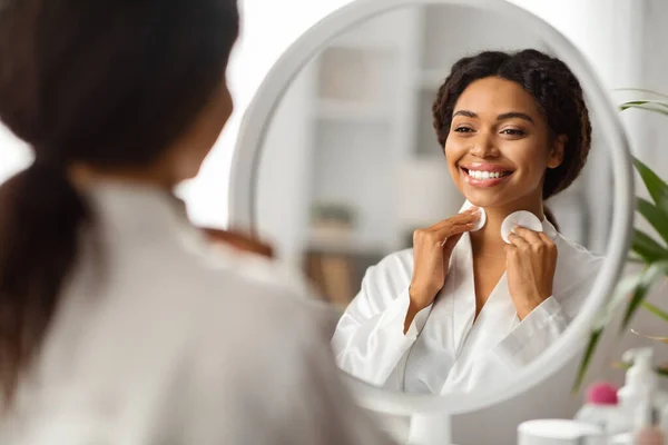 Smiling Black Woman Cleansing Neck Skin With Cotton Pads And Looking In Mirror, Beautiful Young African American Female Making Daily Beauty Routine At Home, Selective Focus On Reflection