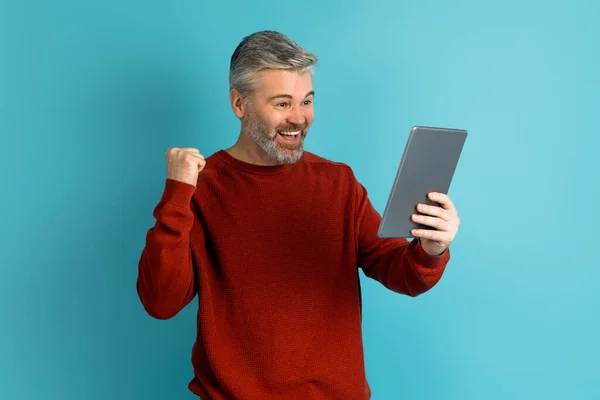 Online bet, gambling, trading. Happy lucky handsome grey-haired middle aged man in red clenching fist, raising hand up, trader celebrating success, holding digital tablet, blue studio background