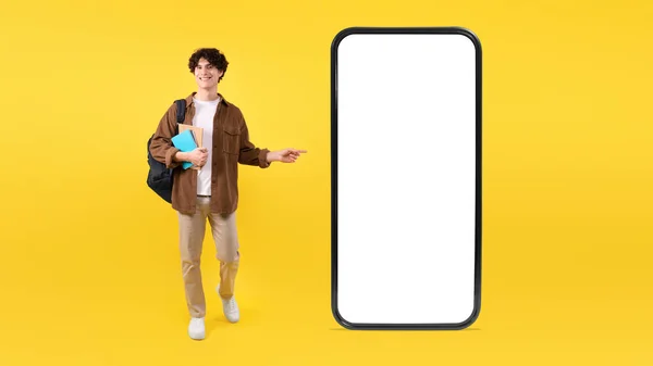 移动报价 年轻的学生用空白屏幕在大型智能手机上推广新应用 用手指指向小工具 背着背包站在黄色背景上 Studio Shot Panorama — 图库照片