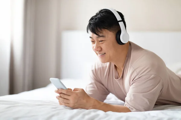 ベッドで携帯電話やワイヤレスステレオヘッドフォンを使用して 家で冷静なパジャマを身に着けている幸せなハンサムな中年のアジア人男性は 音楽を聴き スペースをコピーし 最新の音楽モバイルアプリを楽しんで — ストック写真