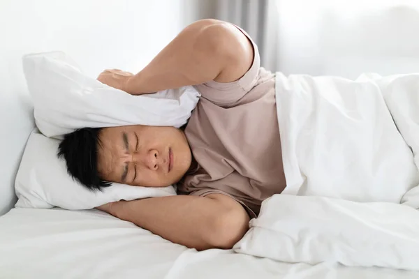 枕で頭をカバーし 家のベッドに横たわっているパジャマを身に着けている激怒した眠れない中年の韓国人男性は 外からのパートナーのいびきや騒音のために眠ることができない クローズアップ — ストック写真