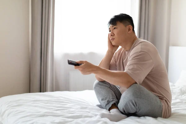 Unhappy bored middle aged asian man wearing pajamas sitting on bed at home, holding remote control and touching his face, watching TV, looking for nice movie or TV show, copy space