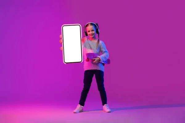 用无线耳机 手持滑板 在霓虹灯下展示巨大的带有白色空白屏幕的手机 色彩艳丽的背景 这是个甜美的 面带微笑的学龄女孩 有着两个辫子 — 图库照片