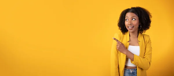 Excited happy black lady in casual outfit posing on yellow background and pointing aside at copy space, panorama. Young african american woman showing nice offer