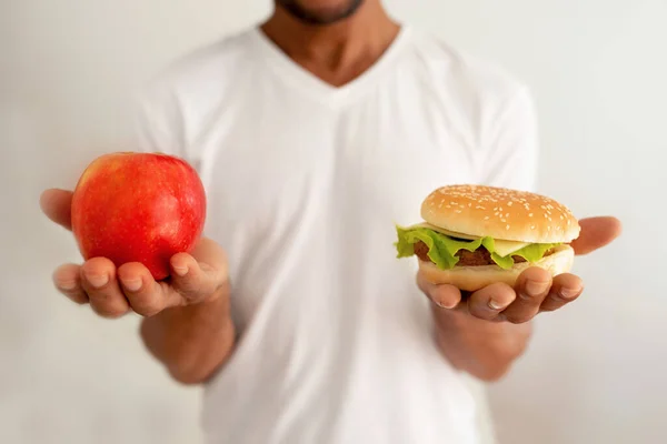 健康食品やジャンクフード 白い壁の背景に対して健康な対不健康な食品の間でバーガーとアップルフルーツを保持黒人男性の作物のショット 不正行為の食事と食事の概念 — ストック写真