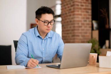 Odaklanmış Asyalı orta yaşlı adam dizüstü bilgisayar kullanıyor, not alıyor, iş raporu yazıyor, evde oturuyor. Çevrimiçi çalışan girişimci ya da serbest çalışan erkek