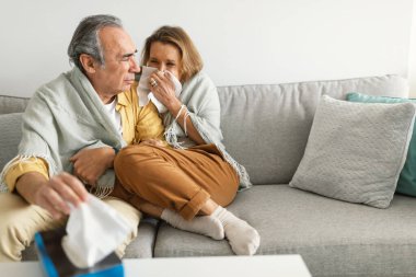 Yaşlı çift soğuktan kıvranıyor, battaniyeye sarılmış oturuyor, burunlarını siliyor ve hapşırıyor, adam peçete alıyor, evdeki kanepede oturuyor, boş yer var.