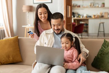 Aile İnternet Alışverişi. Neşeli Japon ebeveynler ve küçük kızı dizüstü bilgisayar ve kredi kartı kullanıyorlar ve ödemeyi evde birlikte yapıyorlar. Ticaret Teklifi ve Banka Hizmeti Reklamları