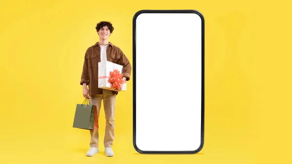 提供网上购物服务 笑的家伙靠近大型智能手机与空白屏风持有包装礼品盒和购物袋 广告应用程序投在黄色工作室背景 — 图库照片