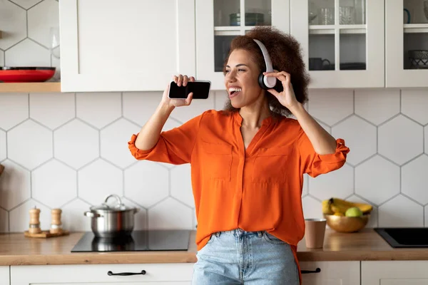 快乐的黑人女人 带着无线耳机和智能手机在厨房里歌唱 快乐的年轻非洲女人 用手机当话筒 在家里玩的开心 复制空间 — 图库照片