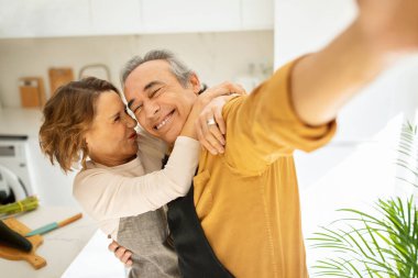 Mutlu, yaşlı, evli bir çift selfie çekiyor, mutfak içinde surat asıyor ve sarılıyor, mutlu anların fotoğraflarını çekerken eğleniyorlar. Modern büyükanne ve büyükbaba ve emeklilik hayatı
