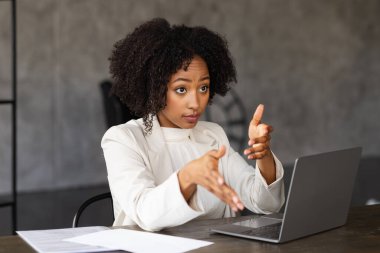 Resmi kıyafet direktörlüğündeki başarılı çekici genç siyahi bayan iş arkadaşları veya çalışanlarıyla brifing veriyor. İş kadını dizüstü bilgisayarın önünde oturuyor, konuşuyor ve el kol hareketi yapıyor.