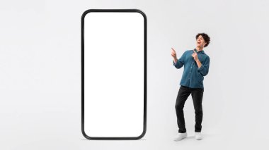 Uygulama Reklamı. Büyük boy akıllı telefonu boş ekran ile işaret eden neşeli adam başvuru önerisi ve beyaz stüdyo geçmişi üzerinden teklif sunuyor. Panorama, Mockup