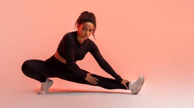 Stüdyo mükemmelliği. Formda bir zenci kadın egzersiz rutininde ustalaşıyor, bacaklarını açıyor ve kameraya bakıyor, şeftali rengi neon arka plan, panorama. Kas antrenmanı, kilo verme konsepti