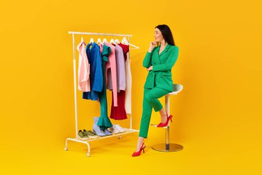 Düşünceli genç bayan elbise raflarına bakıyor ve kıyafet alışverişini seçiyor ve sarı stüdyo arka planında oturan kıyafetler alıyor, tam boy, fotokopi alanı. Moda stilistleri servis kavramı