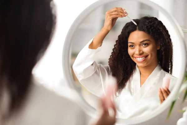 Улыбающаяся Черная Женщина Применяющая Сыворотку Восстановления Волос Стоя Рядом Зеркалом — стоковое фото
