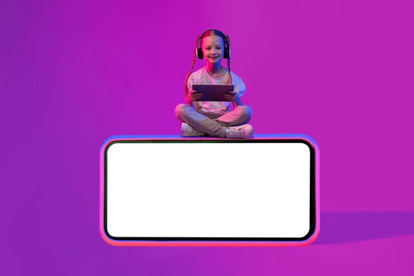 幸せなかわいい子供Preteen女の子で夏服に座って水平電話でホワイトブランクスクリーンモックアップ素敵なオファーのために 使用してデジタルタブレット ワイヤレスヘッドフォンでカラフルな背景にネオンライト — ストック写真