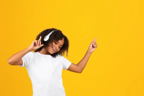 令人高兴的是 身穿白色T恤和无线耳机跳舞 听音乐 享受在黄色工作室背景下独处的年轻非洲女孩 用于放松 有趣的音频应用程序 — 图库照片