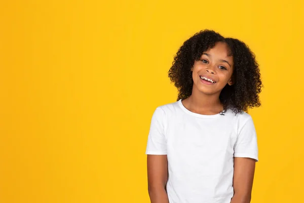 笑着笑着 卷曲的年轻非洲姑娘穿着白色T恤 看着被黄色工作室背景隔离的相机 学生和学校教育 广告和提供 销售和生活方式 — 图库照片