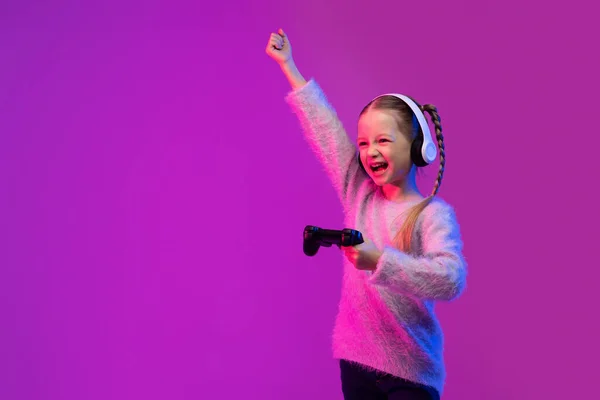 感情甜美的金发少女在霓虹灯下玩彩色背景的电子游戏 用无线耳机和手握操纵杆 举起手 复制空间 儿童的游戏成瘾 — 图库照片