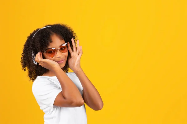 身穿白色T恤 太阳镜和无线耳机的快乐卷曲的非洲裔美国女孩听音乐 享受与黄色工作室背景隔离的闲暇时光 业余时间的放松和快乐 — 图库照片