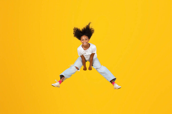 Glad excited curly teen african american girl jumping, freezes in air, has fun on empty space isolated on yellow studio background. Emotions from vacations, education at school, ad and offer