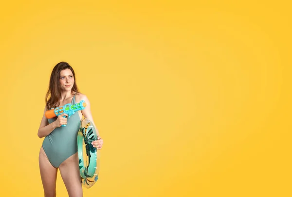 Cheerful pretty woman in swim clothes posing with water gun and inflatable ring, having fun on yellow studio background, free space. Water activity while summer vacation