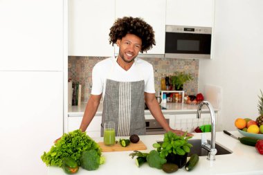 Detoks Tarifleri. Neşeli Siyah Adam Sebze Smoothie İçeceği Pişiriyor veya Yeşil Meyve Suyu Sağlıklı Sebzeler Kullanıyor ve Modern Mutfakta Bitkiler Poz Veriyor, Kameraya Gülümsüyor. Kilo Kaybı Beslenme Konsepti