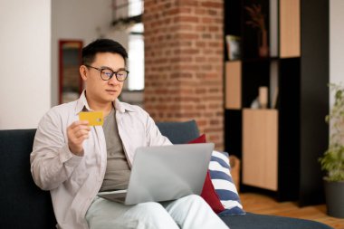 Orta yaşlı Asyalı bir adam modern laptop ve kredi kartıyla koltukta oturuyor, internetten alışveriş yapıyor, evde otururken internetten mal ve hizmetleri ödüyor, yer kopyalıyor.