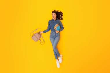 Akademik Joy. Coşkulu Öğrenci Kadın Sırt Çantası ve Çalışan Malzemeler Sarı Stüdyo Arkaplanında Uzanan Kameraya Gülümseyen Tam Uzun Çekim. Modern Üniversite Çalışması Konsepti