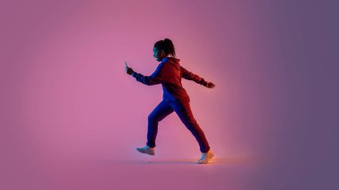 Spor kıyafetli siyahi bir kadın yürüyor ve cep telefonunu pembe neon ışıklı arka planda kullanıyor, tüm vücut uzunluğu, yan görüş, boş alan, afiş.