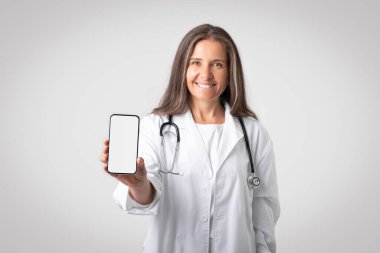 Beyaz önlüklü arkadaş canlısı yaşlı kadın doktor kameraya beyaz ekranlı telefonu gösteriyor, gri arka planda poz veriyor, maketini yapıyor. Profesyonel tavsiye, web sitesi, reklam ve teklif