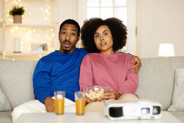 家族の楽しみ 現代のリビングルームの屋内でソファに座っているホームシネマプロジェクターを使用してホラー映画を見て ポップコーンを食べるショックを受けた黒人夫婦 週末のレジャーとエンターテイメント — ストック写真