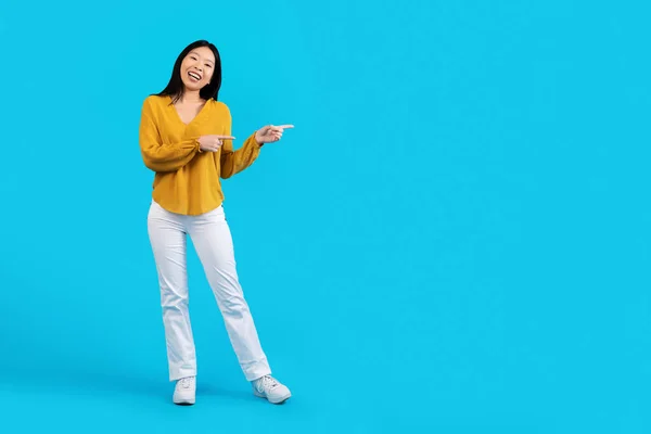 穿着休闲装的快乐 苗条身材的年轻韩国女人 在蓝色的工作室背景上指着广告的复制空间 展示出优美的报价 全身而退 — 图库照片