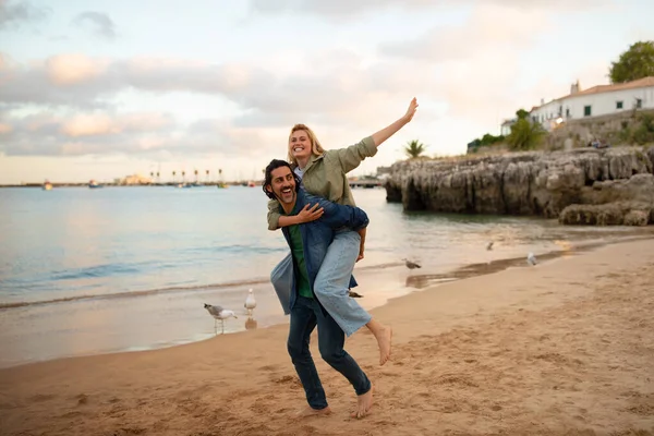 浪漫的年轻夫妇在夕阳西下的沙滩上散步时快乐 男人给女人回旋的车程 快乐的恋人在海滨享受约会 女人回到男友身边 复制空间 — 图库照片