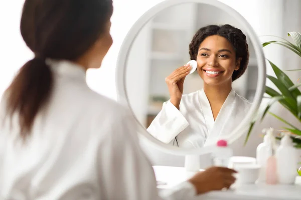 Beautiful African American Woman Cleansing Skin With Cotton Pad Near Mirror At Home, Attractive Black Female Smiling To Her Reflection, Enjoying Daily Skincare Routine, Selective Focus