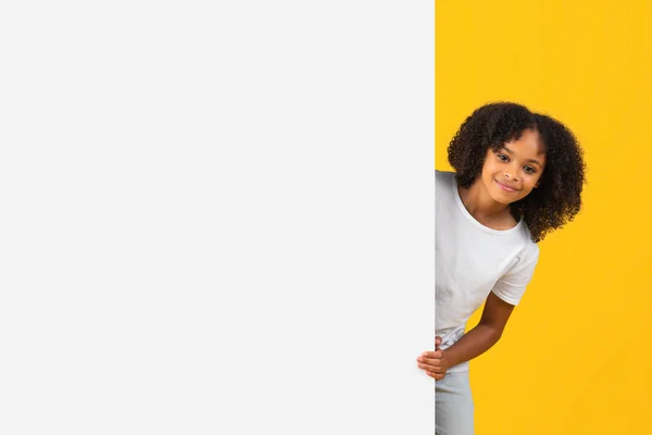 微笑着卷曲的非洲裔美国女孩从巨大的横幅后面看去 空旷的空间被黄色的工作室背景隔开了 学生情绪 学校教育 广告和提供 销售推荐 — 图库照片