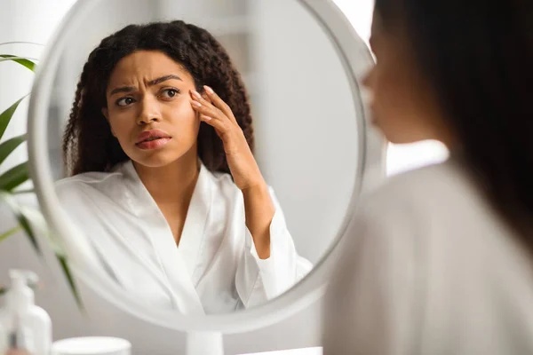 Worried Black Woman Looking In Mirror And Touching Wrinkles On Her Face, Attractive African American Female Examining Fine Lines Near Eyes, Noticed Skin Aging, Selective Focus On Reflection, Closeup