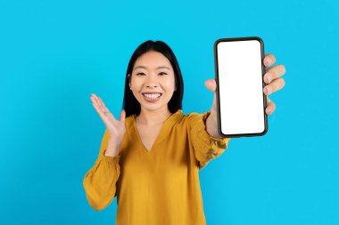 Harika bir online teklif. Duygusal, neşeli, sıradan tişörtlü, genç Koreli bir kadın. Modern cep telefonunu beyaz ekran ve el kol hareketleriyle gösteriyor. Mavi stüdyo arka planında izole edilmiş.