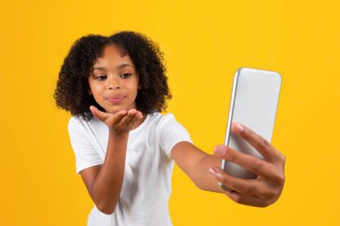 Beyaz tişörtlü mutlu kıvırcık Afrikalı Amerikalı kız akıllı telefondan selfie çekiyor, sarı stüdyo arka planında izole edilmiş öpücük hareketi yapıyor. Çalışma için uygulama, video çağrısı