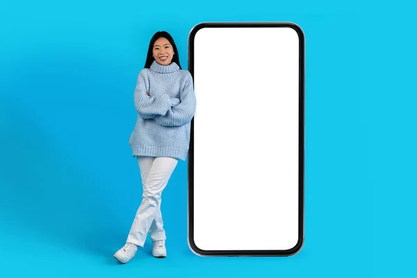 迷人的 快乐的年轻亚洲女人 穿着温暖的毛衣 头戴白色的空屏幕智能手机 模仿漂亮的移动应用程序 在蓝色的工作室背景下隔离 复制空间 — 图库照片