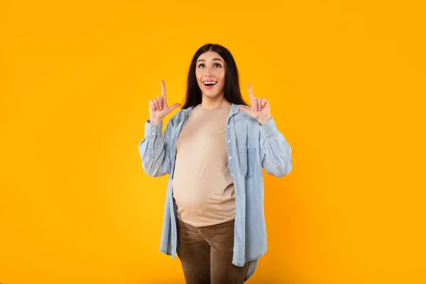 興奮した若い妊娠中の女性は フリースペースに立ち 黄色の背景に立って 期待の女性は彼女の頭の上に広告のための場所を実証 — ストック写真