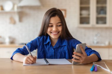 Çevrimiçi Eğitim. Ergenlik çağındaki kız akıllı telefon üzerine ders yapıyor ve evdeki masada otururken notlar alıyor. Mutlu Öğrenci Kız Uzaktan Tutma Telefonu Öğreniyor ve Eğitici Mobil Uygulama Kullanıyor