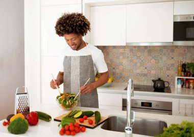 Ev yemeği. Afro-Amerikalı adam yemek yapıyor, salata kasesinde taze sebzeleri karıştırıyor, modern mutfakta tezgahta duran sağlıklı yemek hazırlıyor. İç Tarifler Konsepti