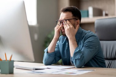 Ofiste dizüstü bilgisayar kullanırken aşırı çalışan genç işadamı, yorgun erkek girişimci masasında oturuyor ve göz kapaklarını ovalıyor, son teslim tarihi stresi yaşıyor,