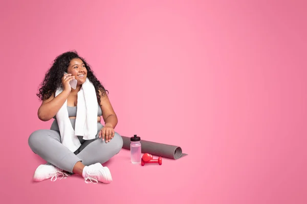 携帯電話で話すスポーツウェアのオーバーサイズの黒人女性は ピンクの背景にダンベルと水のボトルで フリースペースに座っている ワークアウトスポーツのコンセプト — ストック写真
