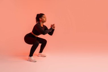 Genç siyahi kadın derin çömelmeler yapıyor ve stüdyoda kırmızı neon arka planda mükemmel bir vücut şekillendiriyor. Motivasyonlu bayan spor yapıyor.