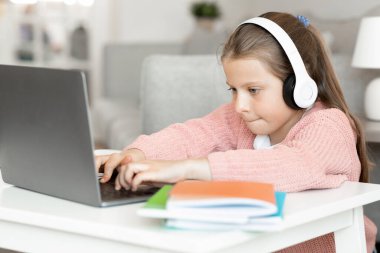 Kulaklık takan, ev ödevi yapan, içerideki bilgisayardan video dersi alan, konsantre olmuş beyaz bir kız. Çocukluk, öğrenme, bilgi ve eğitim, evde cihazla oynanan çevrimiçi oyun