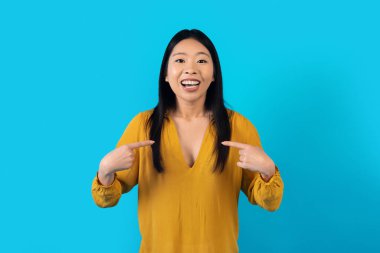 Şanslı, biri. Neşeli, mutlu, pozitif, güzel, Asyalı, sıradan giyinen, kendini işaret eden, mavi stüdyo arka planında izole edilmiş, kopyalama alanı olan, mutlu ve mutlu bir genç kadın.