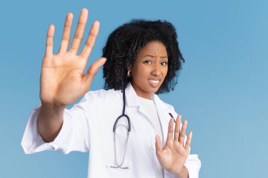 Steteskoplu beyaz önlüklü Afro-Amerikan bayan, mavi stüdyo arka planında soyutlanmış el hareketlerini durduruyor. Tiksinti, kişisel alan, iş yerinde sorunlar
